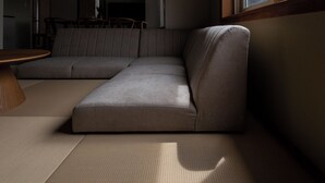 Ein Ecksofa, ein runder Holztisch und eine gewebte Fußmatte in einem Zimmer mit einem Fenster.