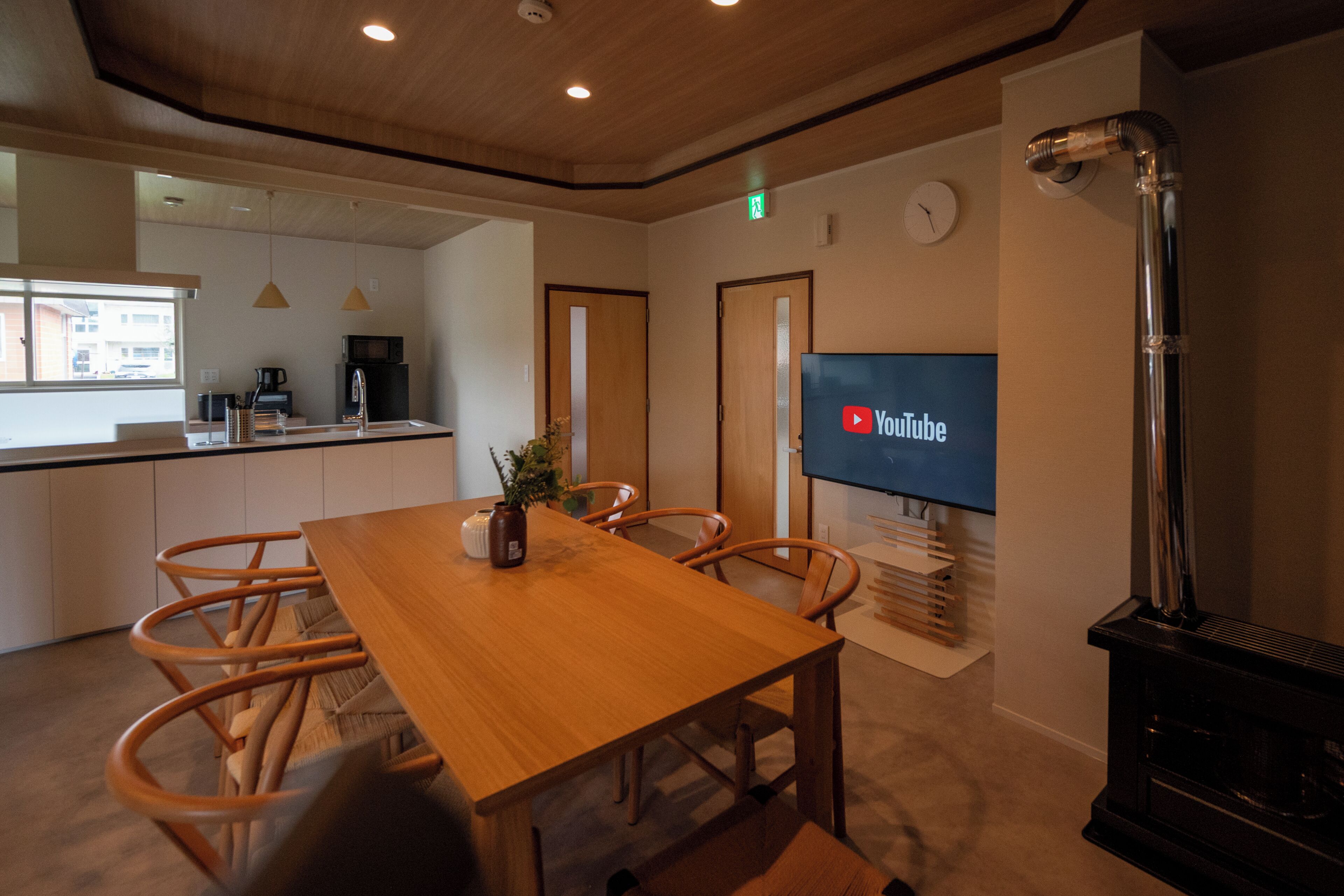 A modern interior with a wooden dining table, chairs, a flat-screen TV, a kitchen area, and a wood-burning stove.