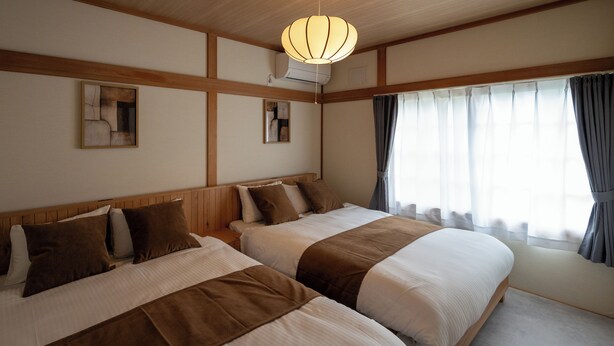 A hotel room with two beds, a large window with curtains, a ceiling fan, and artwork on the walls. - Furano Harmony House (Furano)