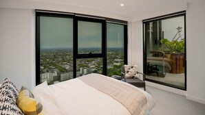 A bedroom with a large bed, a view of a cityscape through floor-to-ceiling windows, and a balcony with a chair and table.