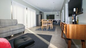 A modern living room with a sofa, ottoman, dining table, chairs, kitchen area, and a TV.