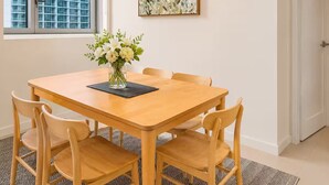 Un comedor con mesa de madera, cuatro sillas, un florero, un cuadro enmarcado y una ventana con vista a un edificio.