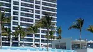 Ein Strand mit weißen Liegestühlen, Palmen und einem modernen Hotelgebäude mit Balkonen.