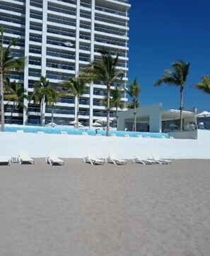 A beach with white lounge chairs, palm trees, and a modern hotel building with balconies. - Harbor 171 | Oceanfront | Infinity Pools | Pet-Friendly (Puerto Vallarta)
