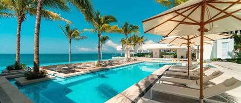 A pool area with lounge chairs and umbrellas, palm trees, and a view of the ocean.