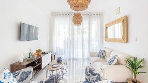 Uma sala de estar moderna com um sofá, uma televisão, uma luminária pendente de vime e uma janela grande com cortinas translúcidas.