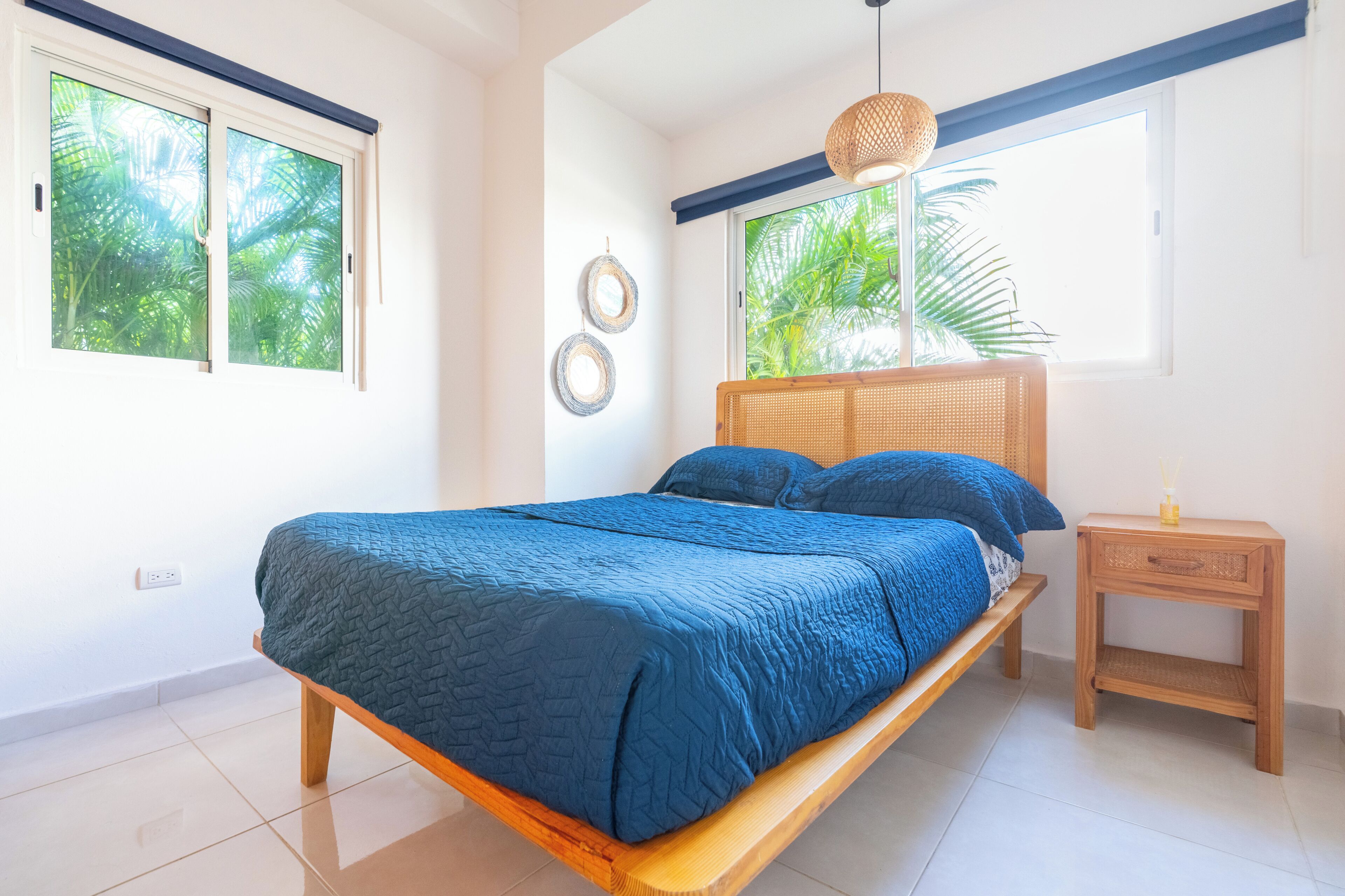 A bedroom with a bed, a nightstand, a window with a view of palm trees, and a ceiling light.