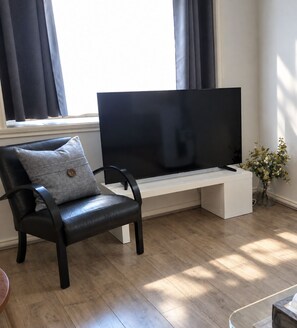A modern living room with a black leather chair, a flat-screen TV on a white stand, and a vase with flowers on the floor. - Park view home (Vancouver)