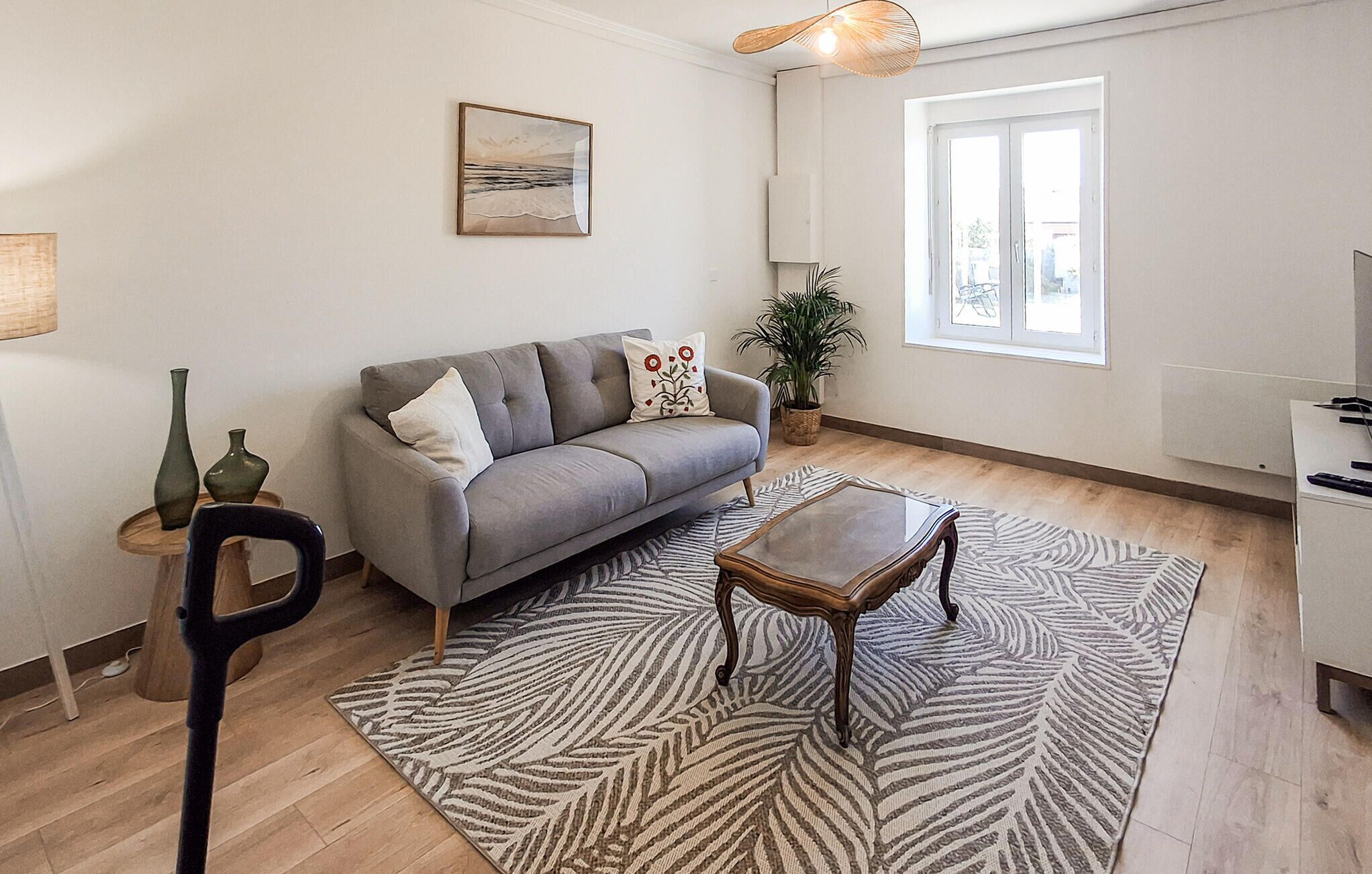Ein Wohnzimmer mit grauem Sofa, Couchtisch aus Holz, gemustertem Teppich, Zimmerpflanze und einem Bild an der Wand.
