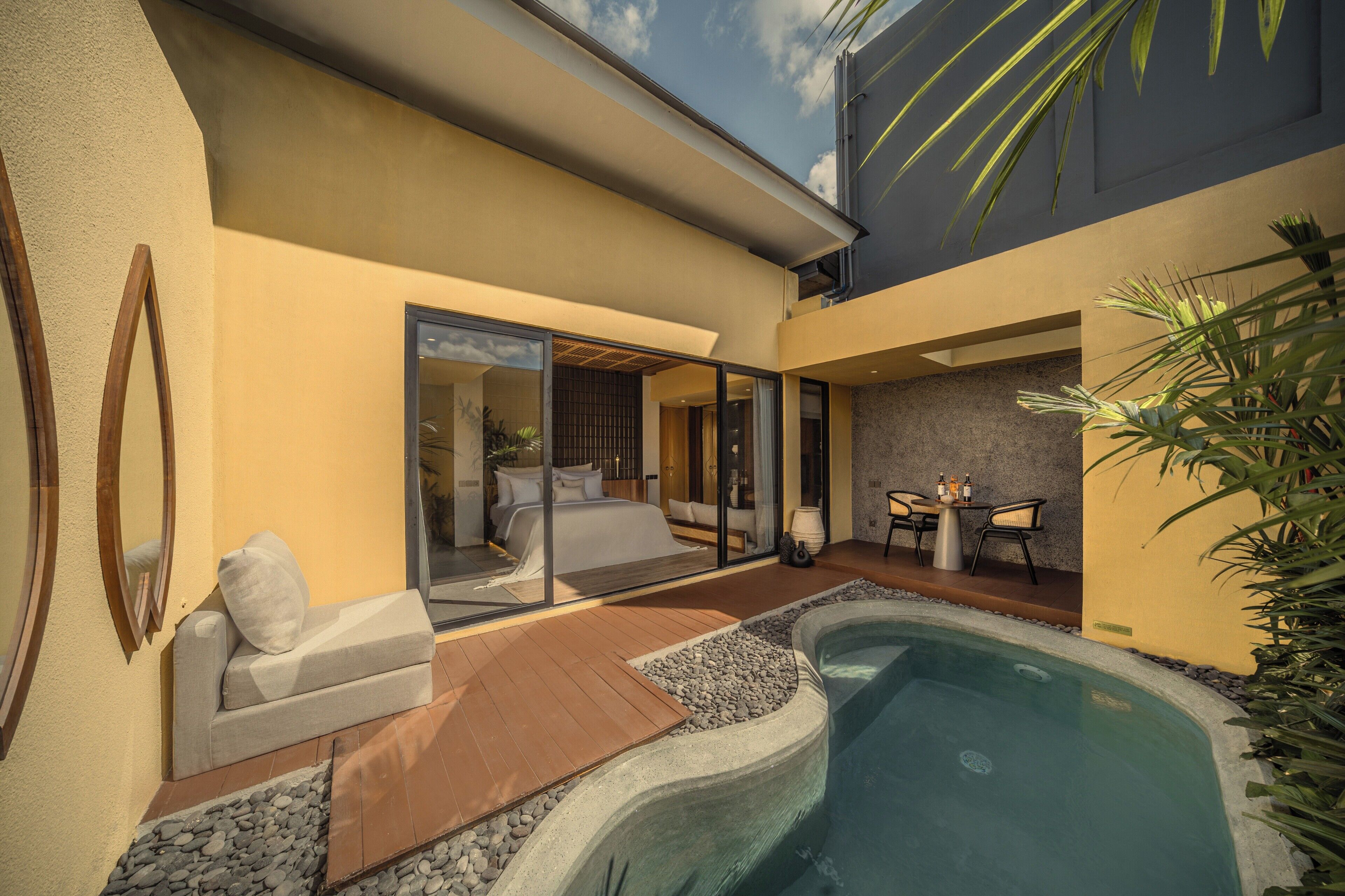 Un área de piscina moderna al aire libre con un jacuzzi, una zona de estar con mesa y sillas, y un ventanal que da a un dormitorio.