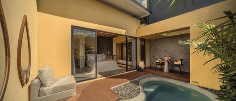 Un área de piscina moderna al aire libre con un jacuzzi, una zona de estar con mesa y sillas, y un ventanal que da a un dormitorio.