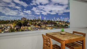 Een balkon met een houten tafel en stoelen, met uitzicht op een stadsgezicht met gebouwen en een heldere lucht.