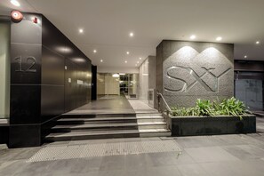 Ein moderner Hauseingang mit einer Treppe zum Empfangsbereich, einem großen 'SX'-Schild und einem Feuermelder an der Wand.