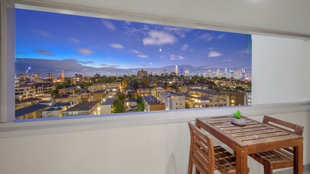 Uitzicht op een stadsgezicht vanaf een balkon met een tafel, stoelen, een plant in pot en een raam met zicht op gebouwen en een kerktoren.