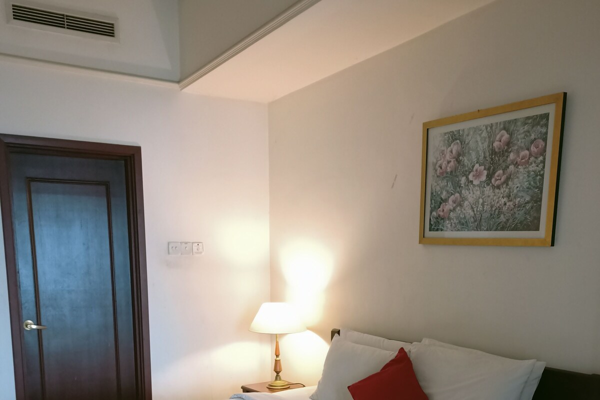 Ein ordentlich bezogenes Bett mit weißer Bettwäsche, einem Holz-Kopfschild, einem roten Kissen und einer Wandlampe.