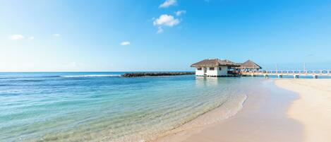 A beach with clear water, a bungalow-style building, and a pier extending into the sea.