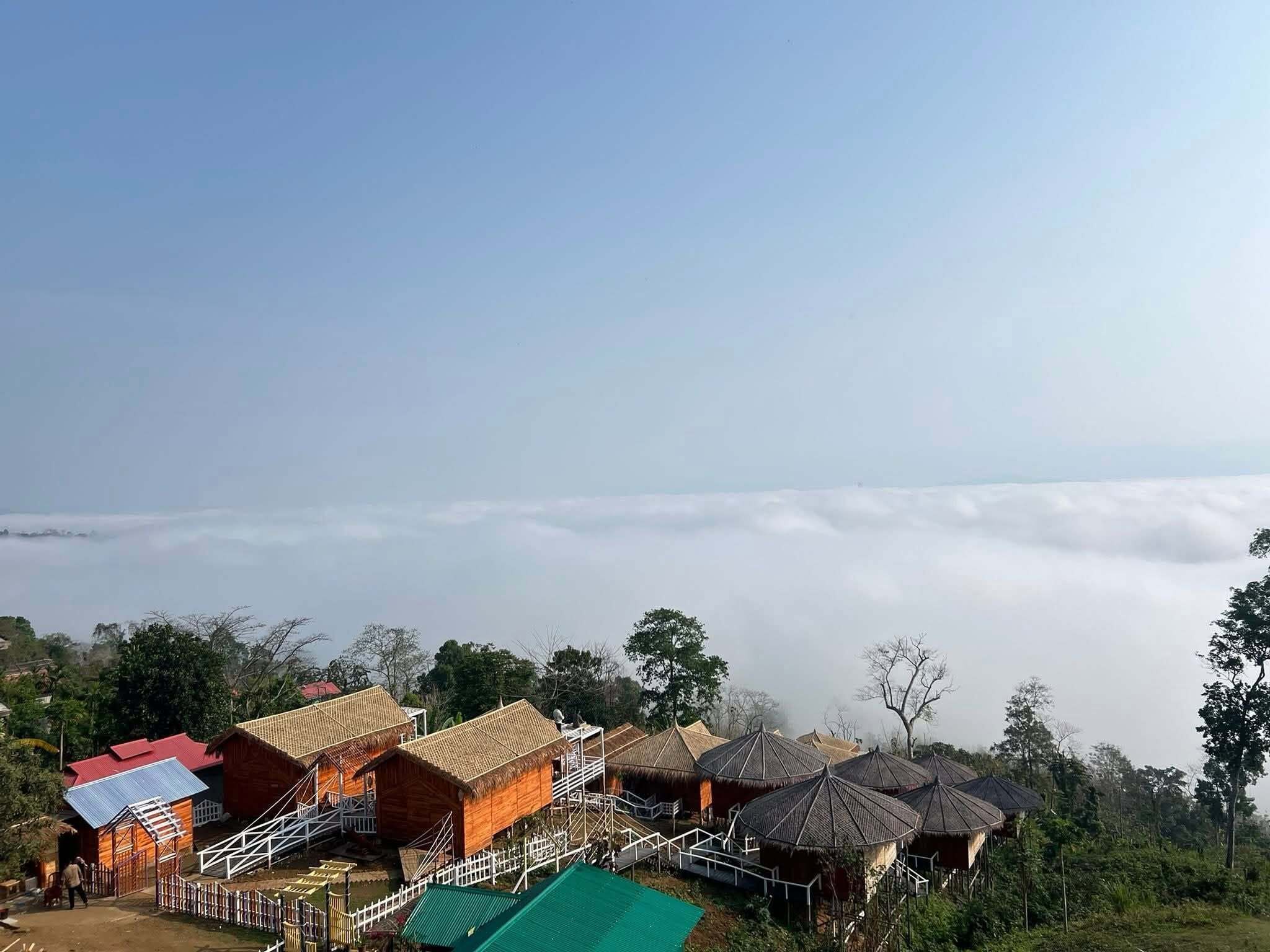 A view of a mountain resort with wooden cabins and thatched-roof huts, surrounded by lush greenery and a sea of clouds below.