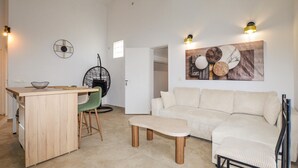 Un espacio habitable moderno con una mesa de comedor de madera, una silla verde, un sofá color beige y un pequeño banco. Hay un reloj de pared y una obra de arte abstracta sobre el sofá.