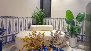 A modern room with a blue accent wall, a white circular vase with yellow flowers, a beige sofa, and a potted plant. - Indigo - Luxury Bathtub Studio at Golf Course Rd (Gurugram)