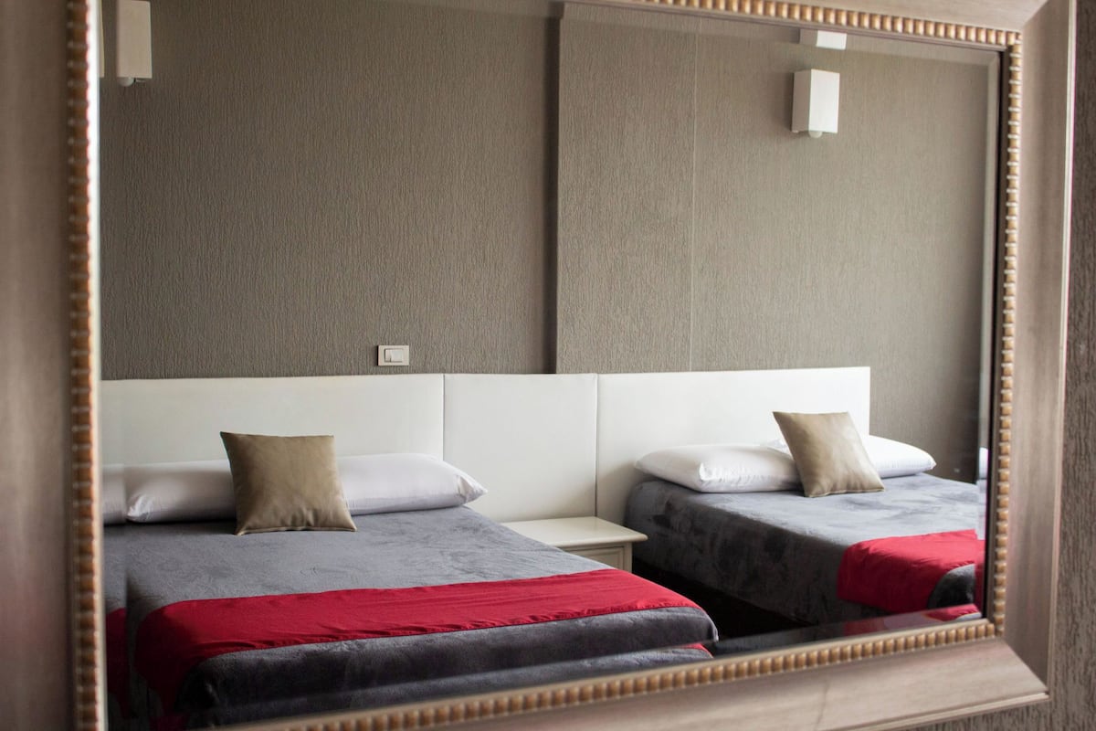 A mirrored view of a hotel room with two beds, a white headboard, and a grey textured wall.
