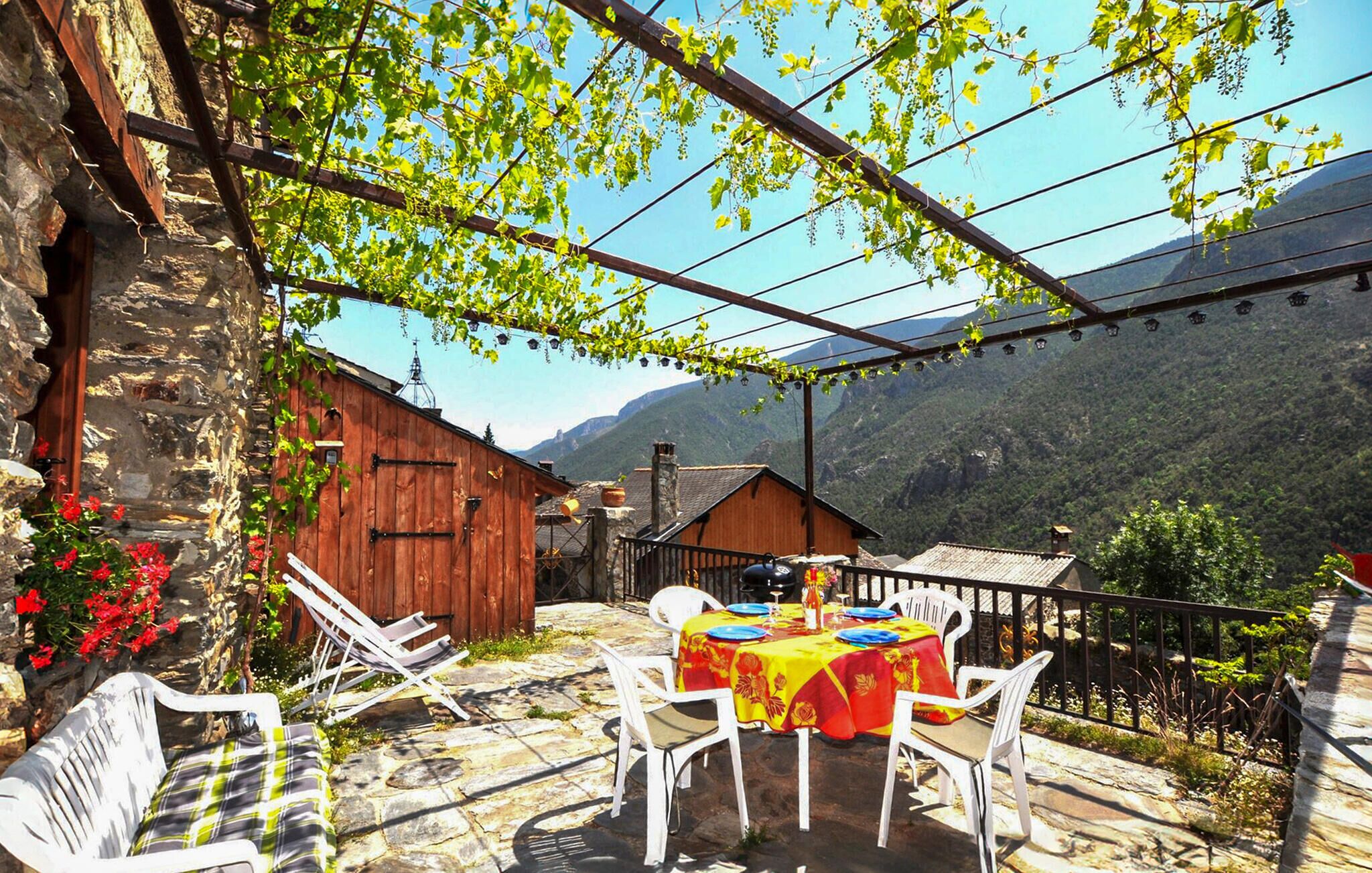 A stone building with a wooden door, a table with a red tablecloth, white chairs, and a view of mountains.