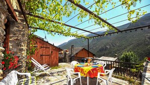 A stone building with a wooden door, a table with a red tablecloth, white chairs, and a view of mountains. - Authentique maison Montagne (Nohèdes)