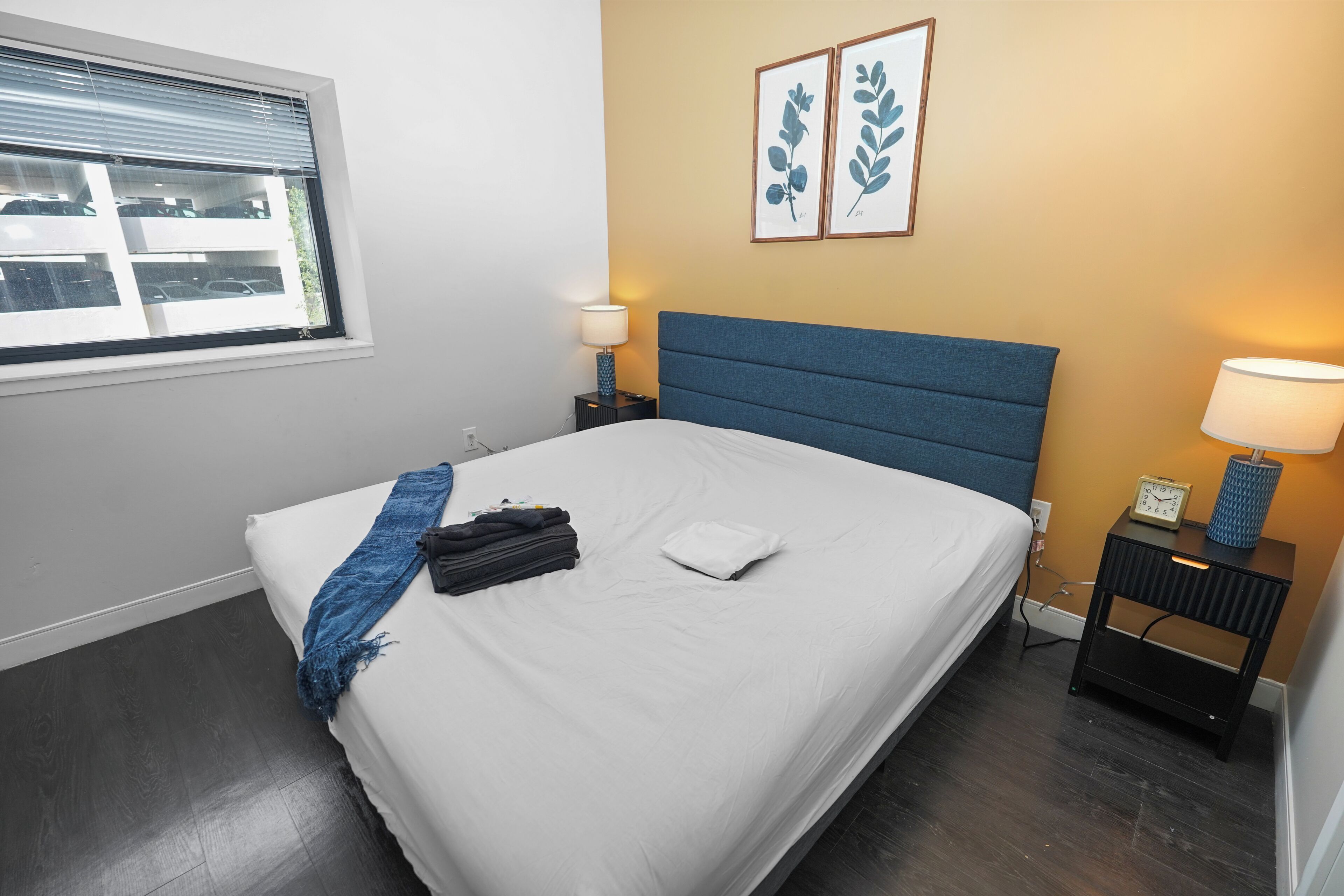 A neatly made bed with a blue headboard, a window with blinds, two bedside lamps, and a wall-mounted clock.