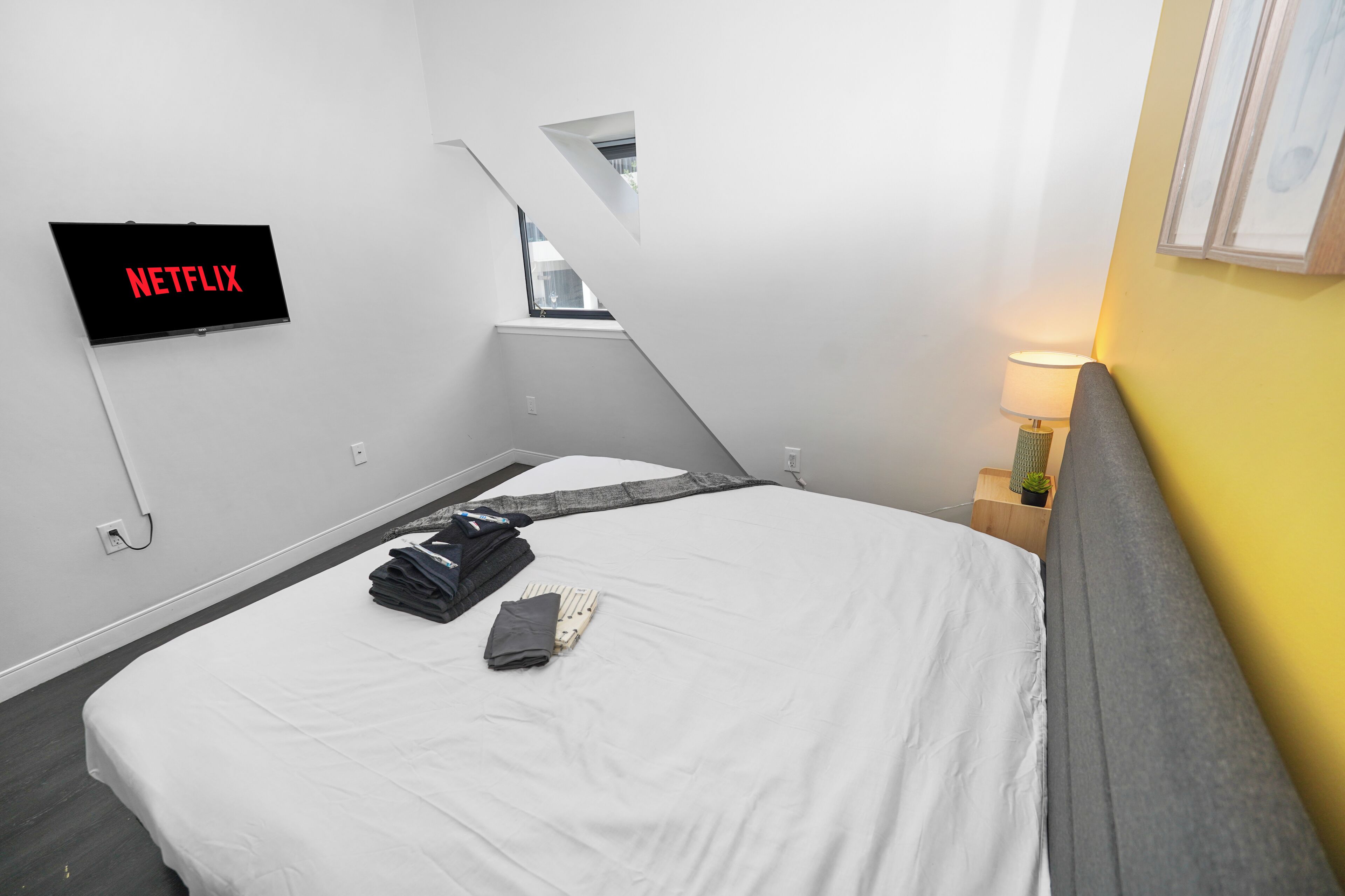 A hotel room with a bed, a TV mounted on the wall, a bedside lamp, and a window.