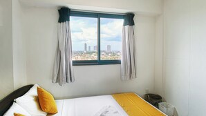 Ein Hotelzimmer mit Bett, Fenster mit Vorhängen und Blick auf die Stadt sowie einer kleinen Mülltonne.
