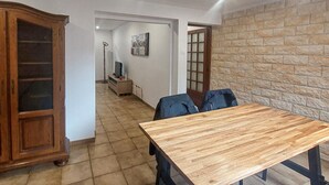 A hallway with a wooden table and chairs, a television on a stand, and a stone wall. - Jolie maison avec cour (Grand-Fort-Philippe)