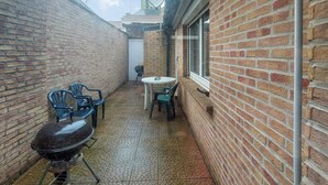 A narrow outdoor patio with a brick wall, a small round table, and two chairs. - Jolie maison avec cour (Grand-Fort-Philippe)