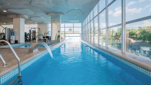 An indoor swimming pool with clear glass walls, a view of the outdoors, and exercise equipment in the background. - Corporate Stays Miami (Asunción)
