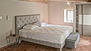 Un dormitorio con cabecera capitonada, cama individual, mesita de noche con lámpara, un banco y un armario.