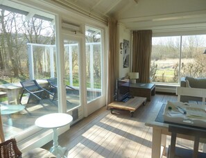 A spacious room with large windows, a television, a sofa, and a dining table. - Bungalow Near Dishoek Beach (Koudekerke)