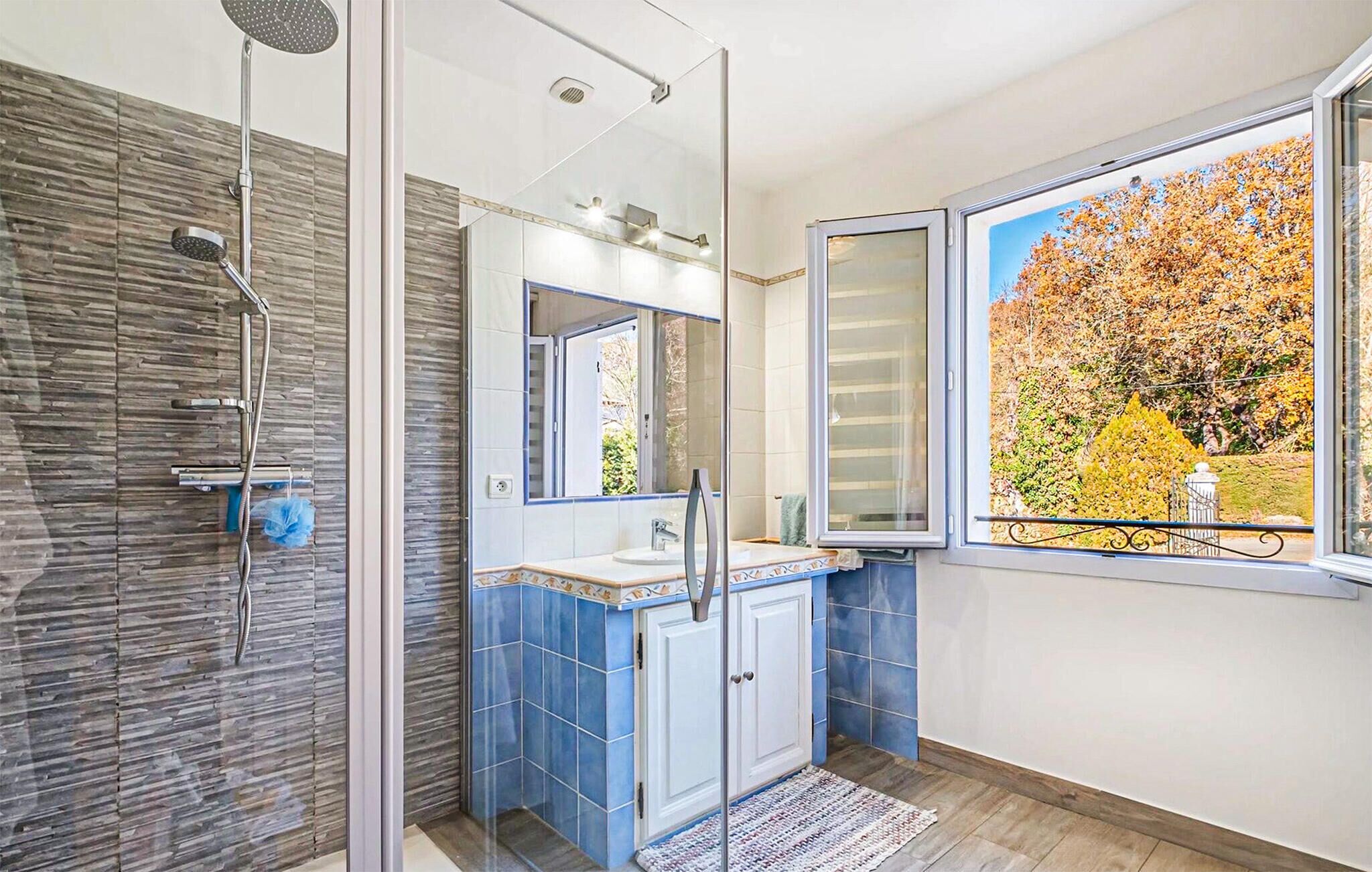 A modern bathroom with a glass shower enclosure, a large mirror, and a view of autumn foliage through a window.