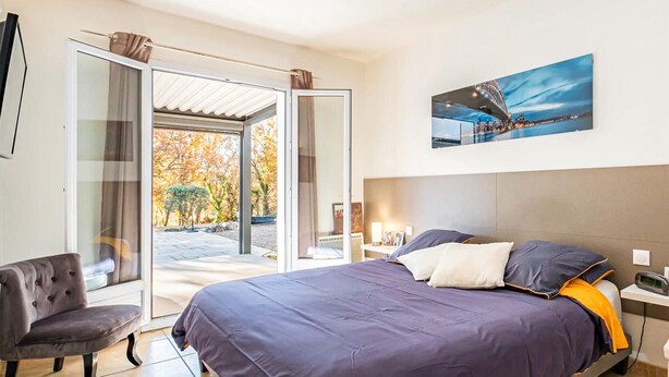 Quarto com cama, uma cadeira, um quadro e vista através de uma porta de vidro.