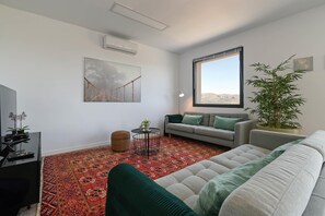Ein Wohnzimmer mit Sofa, Couchtisch, Fernseher, einem gemusterten Teppich, einem Gemälde und einem Fenster mit Ausblick.