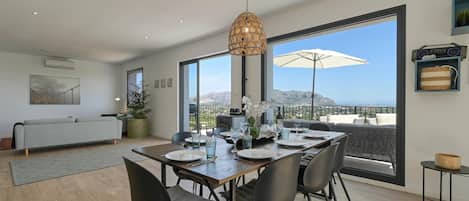A modern dining area with a wooden table set for four, surrounded by chairs, a hanging light fixture, and a large window offering a view of the mountains.