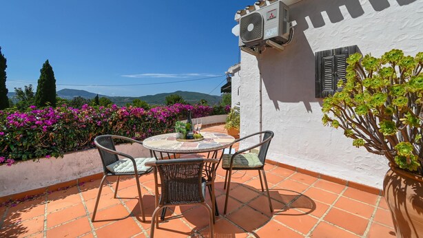 Um terraço com mesa e cadeiras, um vaso de planta e uma parede branca com janela e unidade de ar condicionado.