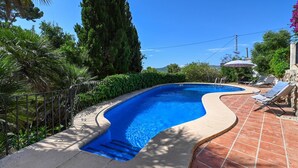 Uma piscina com borda curva, rodeada por um pátio ladrilhado e vegetação.