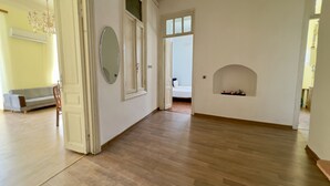 A room with wooden flooring, a white door, a mirror, a fireplace, and a bed in the background.