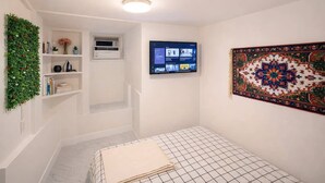 A bedroom with a bed, a mounted TV, a wall-mounted air conditioner, a shelf with books and a potted plant, and a decorative tapestry.