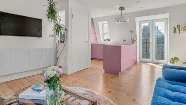 Een moderne woonkamer met een blauwe bank, een houten vloer, een roze keukeneiland en een groot raam.