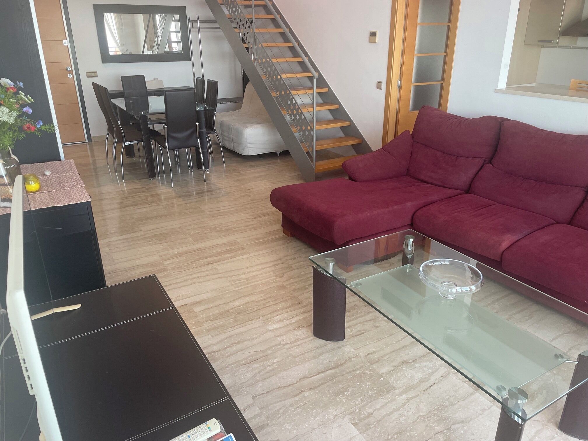 Una sala de estar con una mesa de centro de vidrio, un sofá rojo, un comedor con sillas y una escalera.