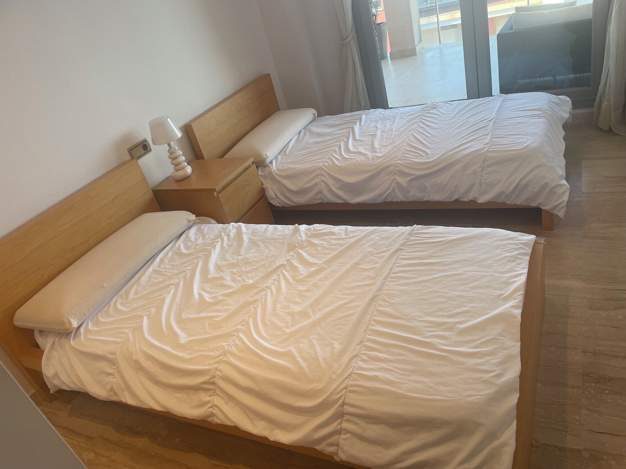 A hotel room with two beds, a wooden headboard, a nightstand with a lamp, and a view of the outside through a large window.