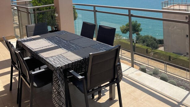 En balkon med bord og stole, der kigger ud over et klart blåt hav og fjerne kyster.