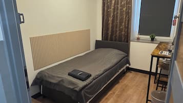 Quarto compacto com cama, uma mesinha, janela com cortinas, radiador e um quadro fixado na parede.