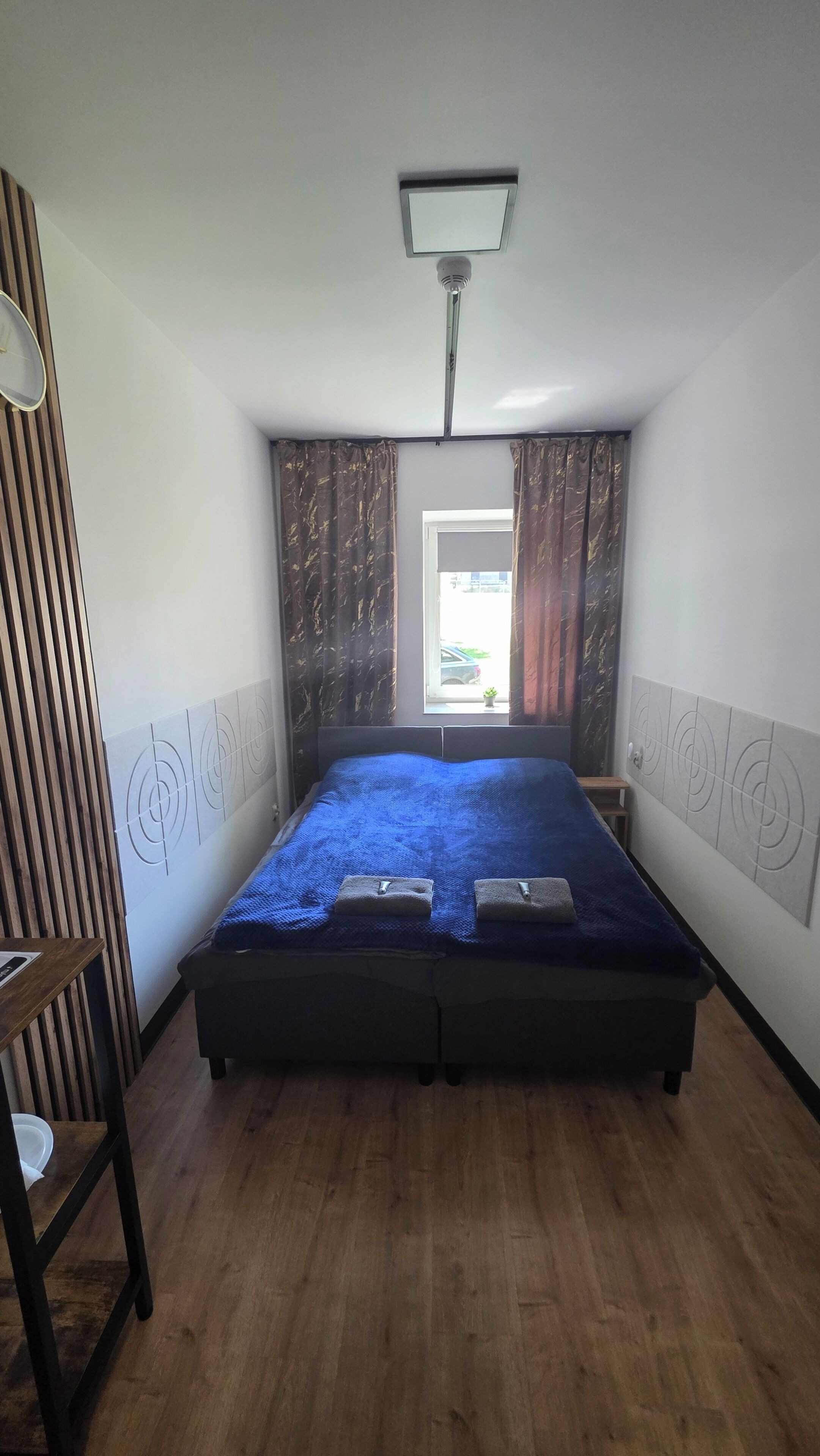 Un dormitorio con cama, ventana con cortinas, luz de techo y suelo de madera.