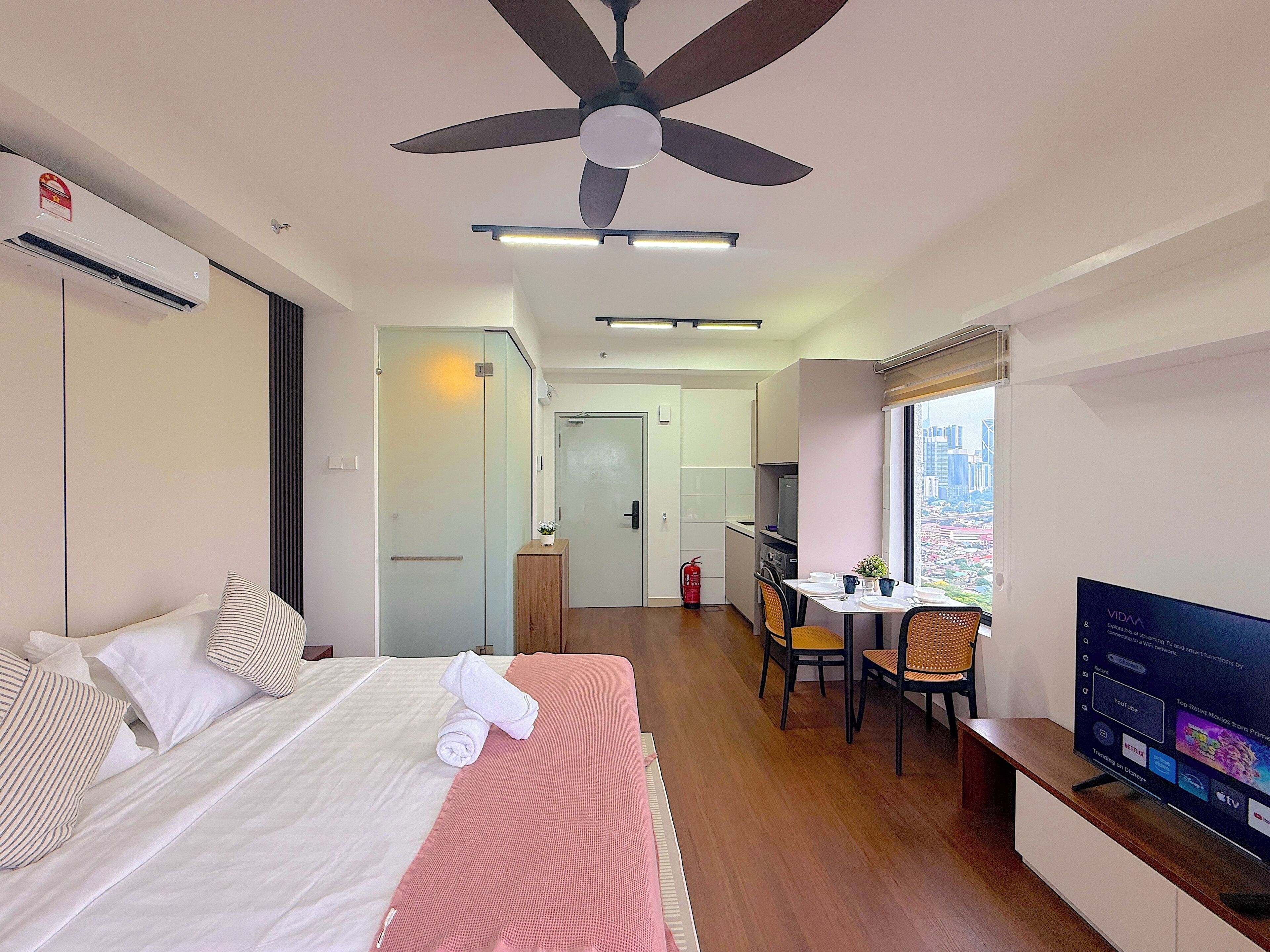 A hotel room with a bed, a ceiling fan, a TV, a dining area with a table and chairs, and a view of the city through a window.