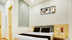 A hotel room with a bed, bedside table, lamp, and a window with blinds. - Saver Single House of Dreams (Phagwara)